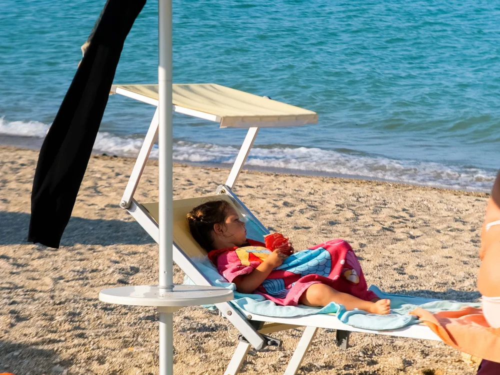 Mädchen liegt auf Strandliege mit buntem Handtuch und Spielsachen im Sand