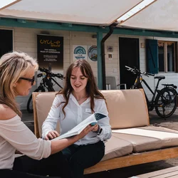 Due donne sedute su un divano all'aperto consultano un catalogo davanti a un negozio di biciclette