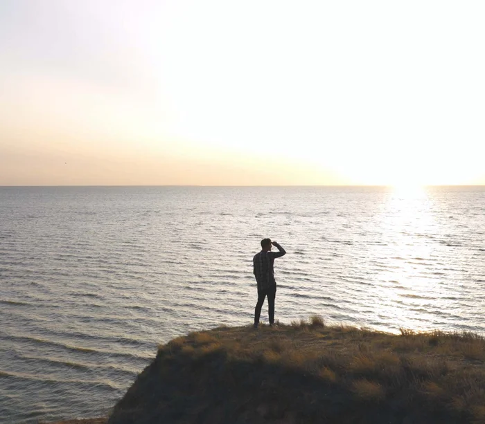 Person steht auf einer Klippe und blickt in den Sonnenuntergang über dem Meer