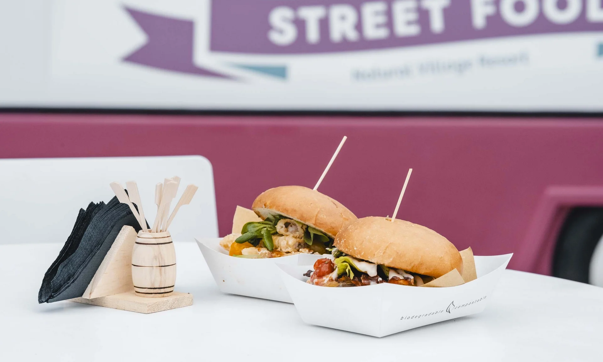 Zwei Gourmet-Burger in Papierschalen auf weißem Tisch mit Servietten und Foodtruck im Hintergrund