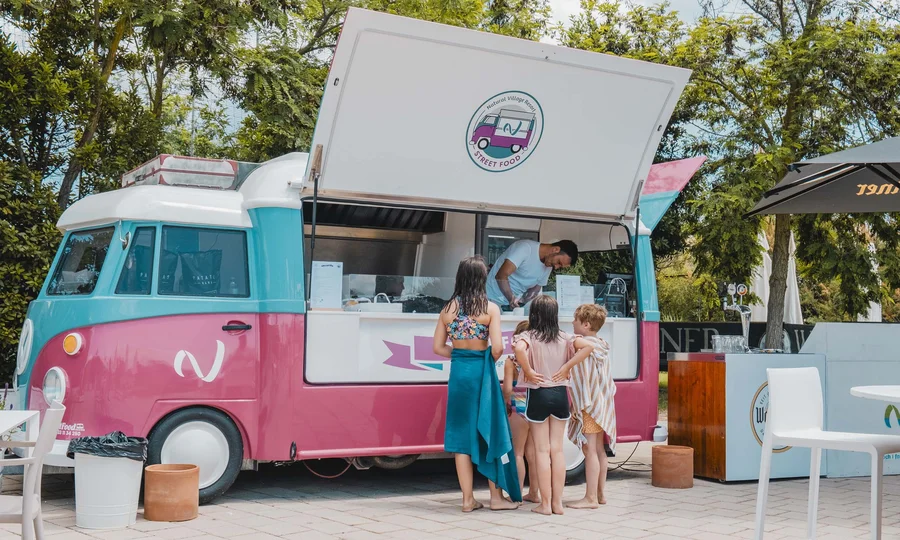 Kinder in Badebekleidung bestellen an rosa-blauem Foodtruck draußen neben Tischen