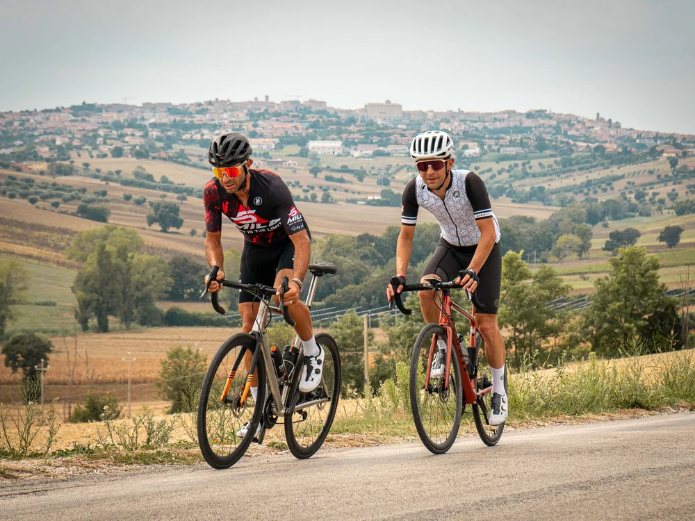 Due ciclisti in abbigliamento da corsa pedalano su strada di campagna con paese sullo sfondo