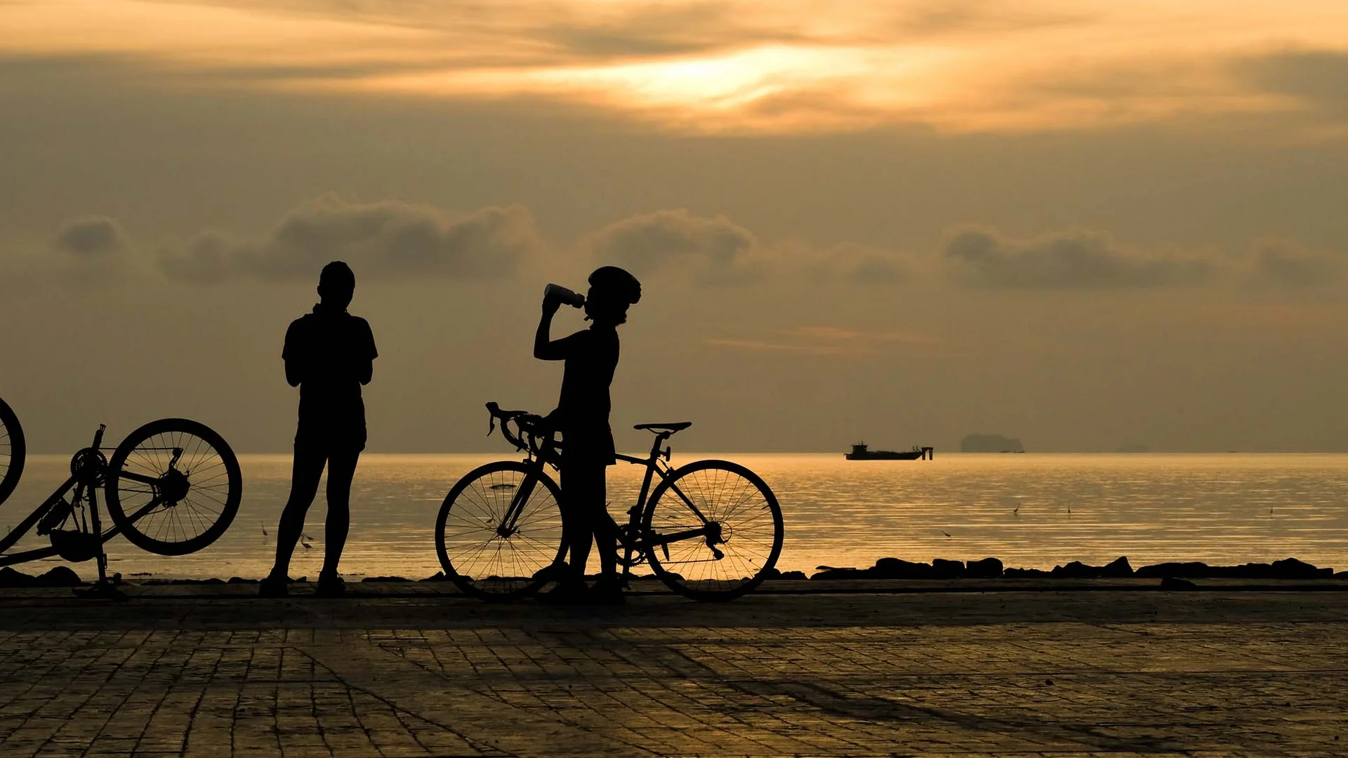 Zwei Radfahrer mit Fahrrädern bei Sonnenuntergang am Meer, einer trinkt Wasser
