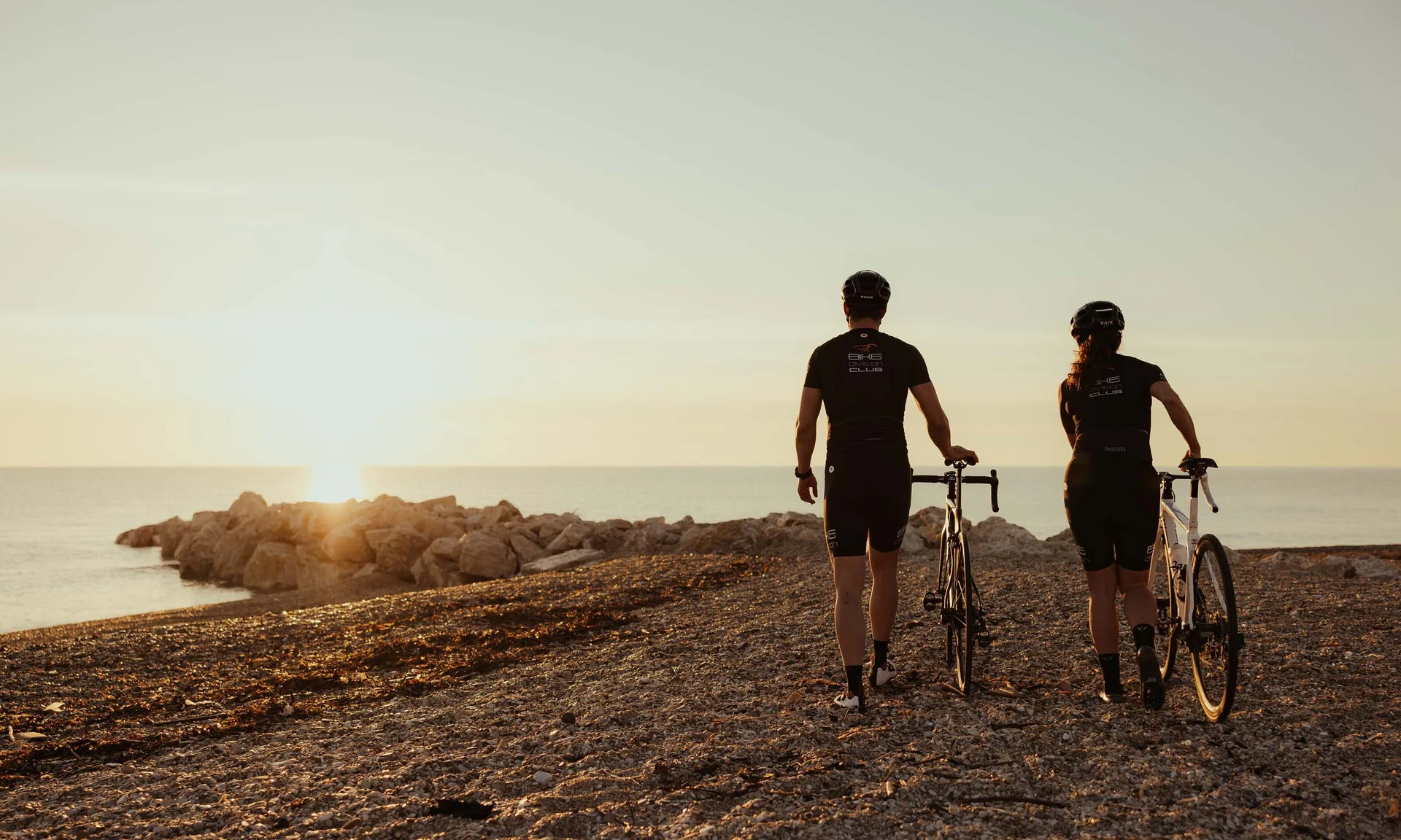 Zwei Radfahrer mit Fahrrädern am Kiesstrand bei Sonnenuntergang